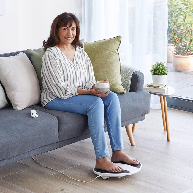 Image of a woman sat in her living room on the sofa with a mug and her feet on the prohealth device 