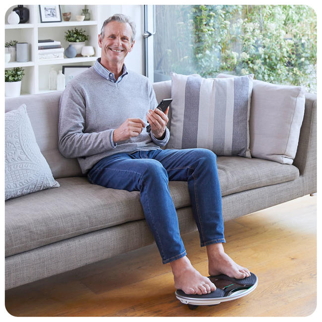 Image of a man in his living room sitting on the sofa with his feet on the deivce and holding his phone 