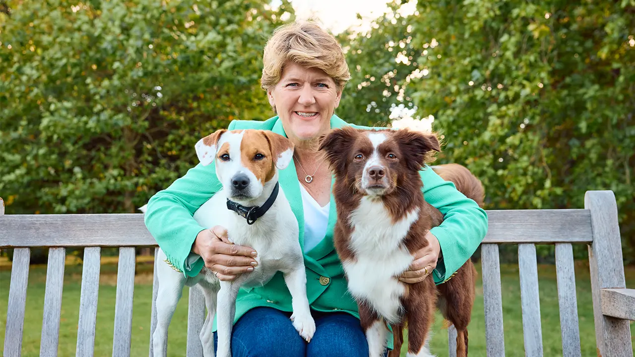 Clare Balding outside sat on a bench with dogs either side of her 