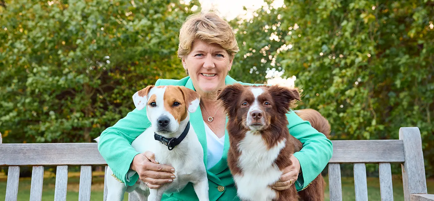 Clare Balding outside sat on a bench with dogs either side of her 
