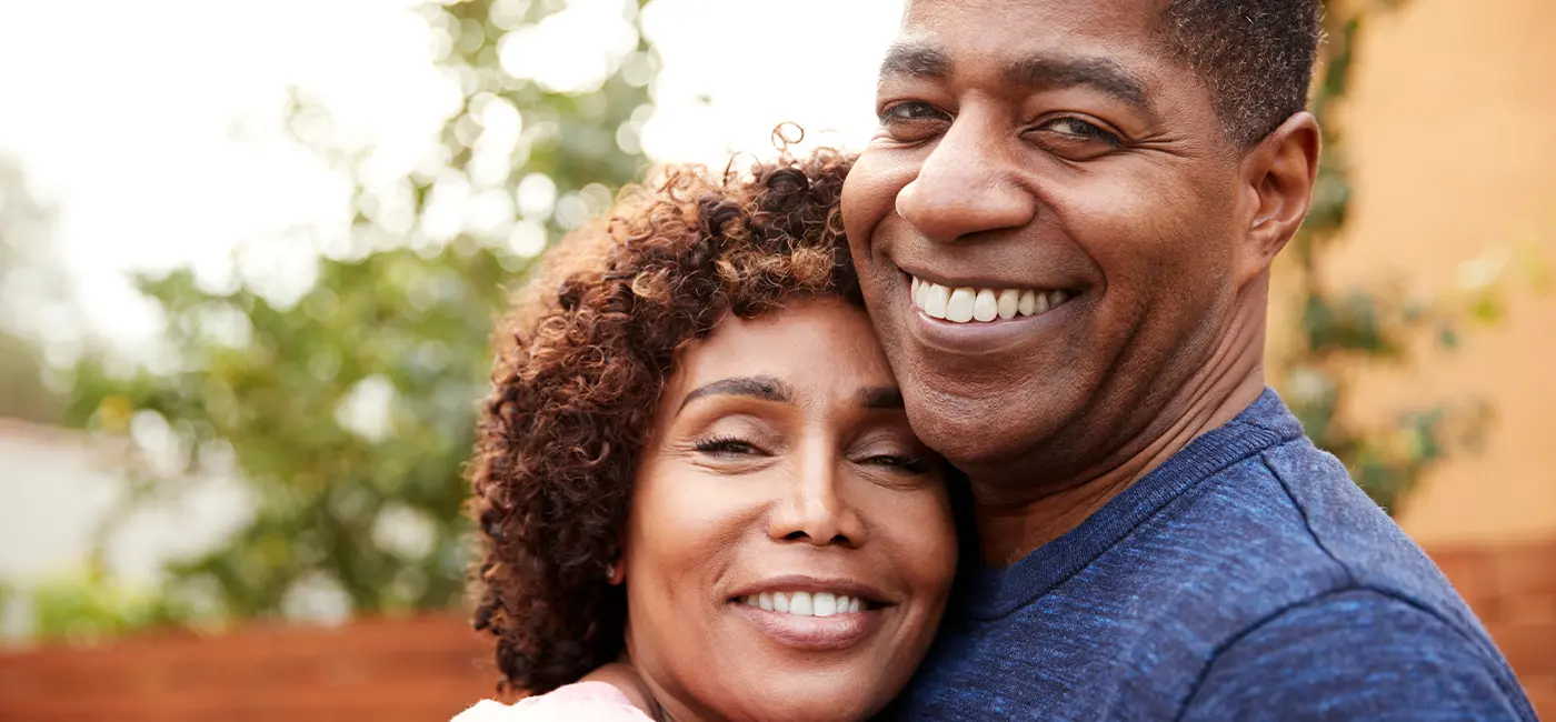 A headshot of a middle-aged couple who hold each other in an embrace. They smile at the camera.