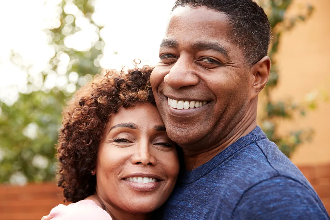 A headshot of a middle-aged couple who hold each other in an embrace. They smile at the camera.