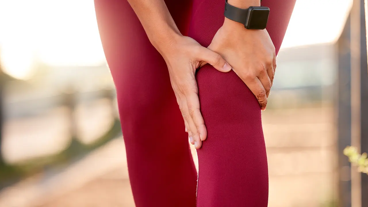 Close-up of a woman wearing burgundy leggings and holding her knee joint with both hands.