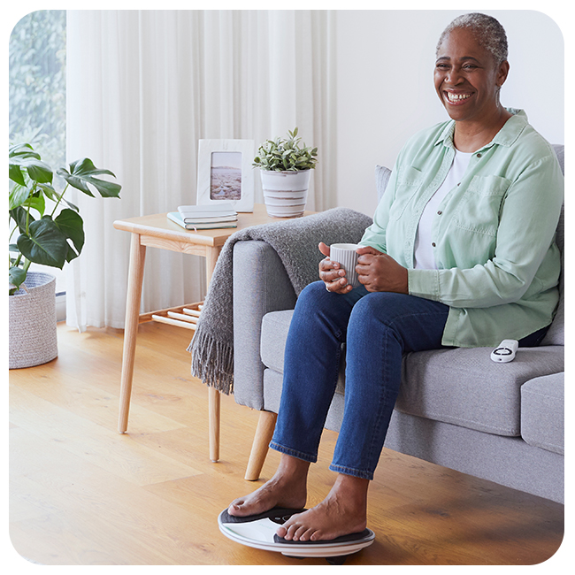 Woman sat on the sofa smiling, with her feet on a medic device 