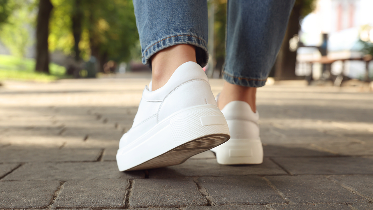 Person, die auf einem von Bäumen gesäumten Weg steht und von der Kamera wegschaut, trägt Jeans und ein Paar weiße Turnschuhe