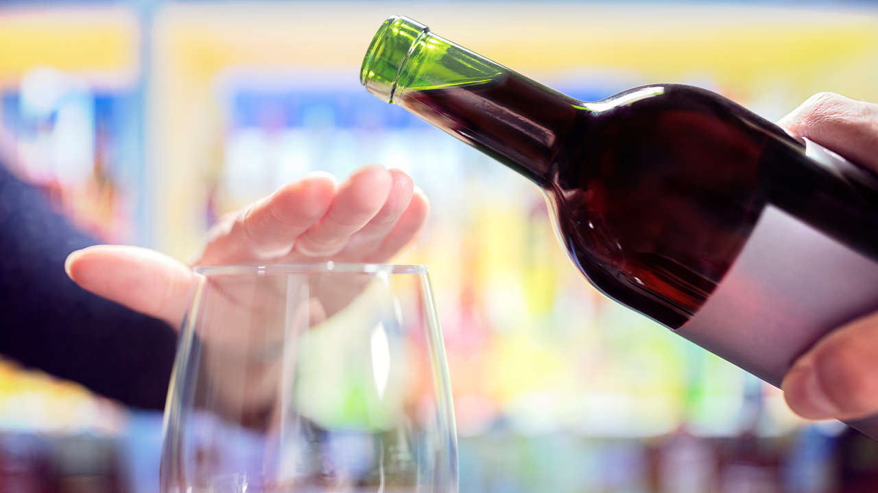 Image of a glass and a bottle of wine, with someone putting their hand over the glass