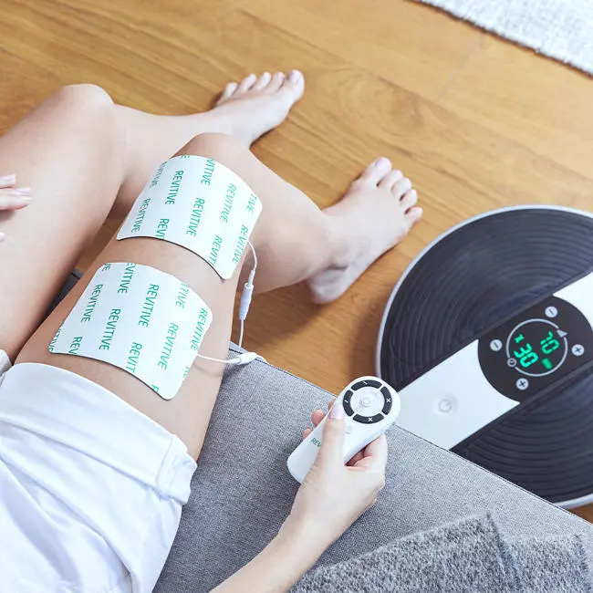 Image of women with the thigh pads on her thighs with the device next to her feet with remote in her hand