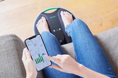  Close up of a womans legs on the coach device, holding her phone with the app open ready to use