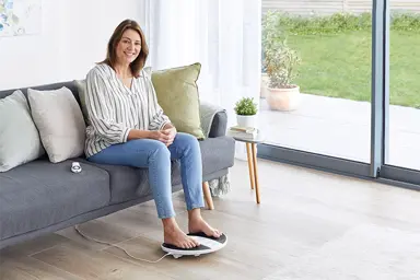 Image of woman in her living room sitting on her sofa with her feet on the prohealth device 